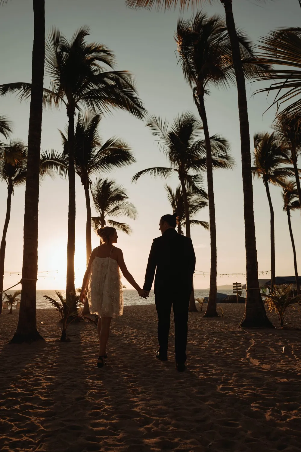 The couple walking away through palms as the sun goes down
